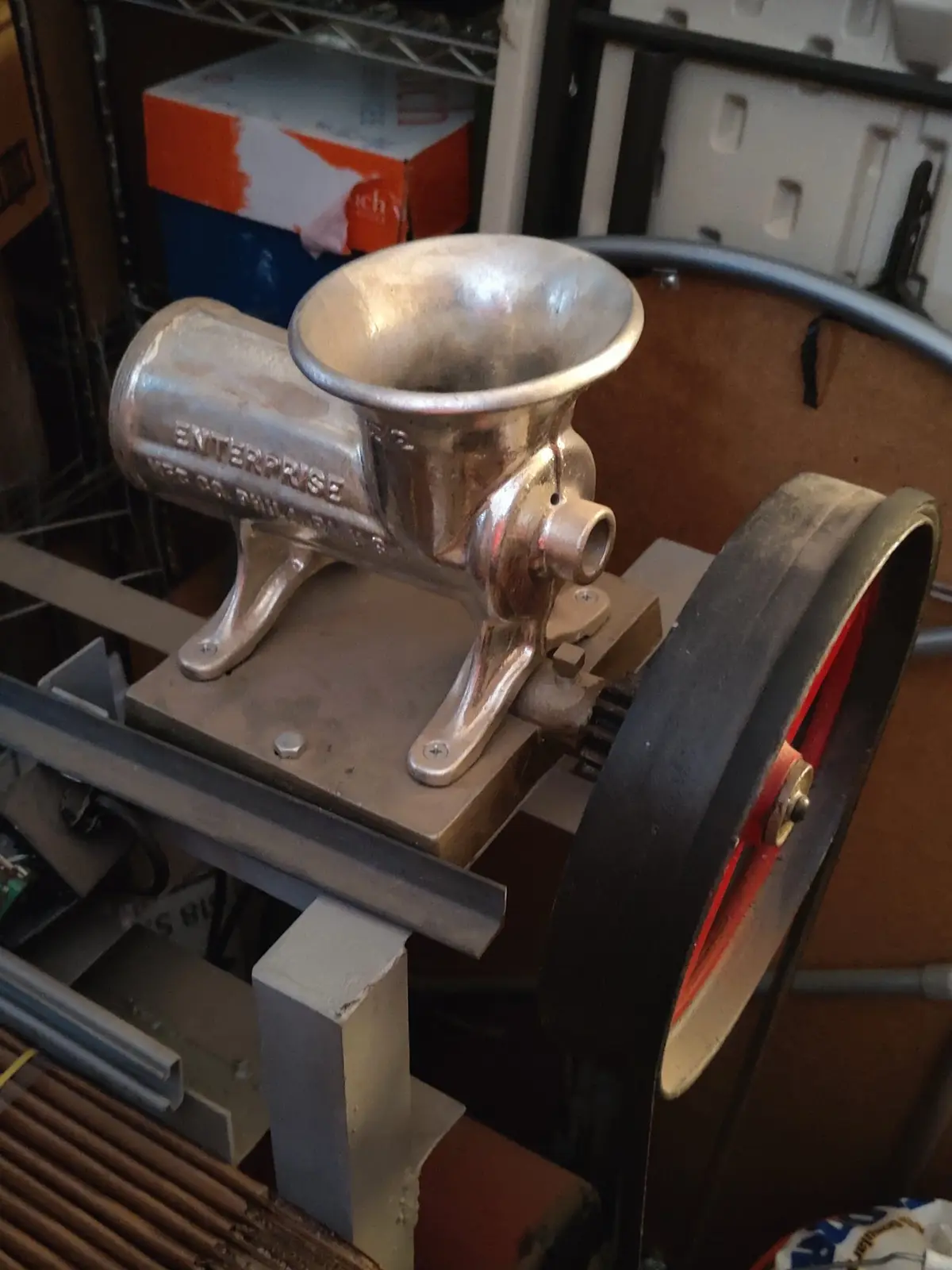 A vintage silver meat grinder branded 'ENTERPRISE' sits on a metal stand with a large black flywheel, surrounded by workshop…