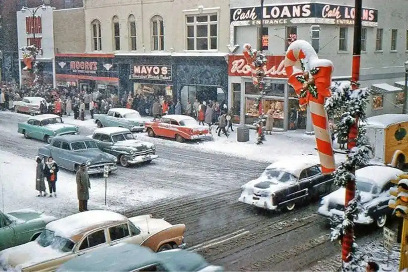 A vintage snowy street scene with classic 1950s cars and pedestrians, featuring storefronts like 'Moont's' and 'Mayo's', and…