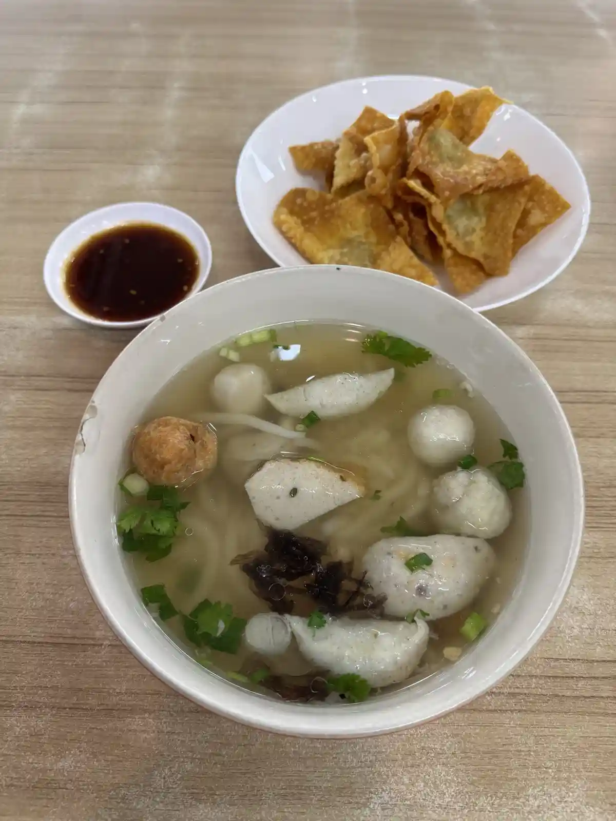 A top-down photo shows a white bowl of fishball noodle soup with various dumplings, noodles, and herbs in clear broth…