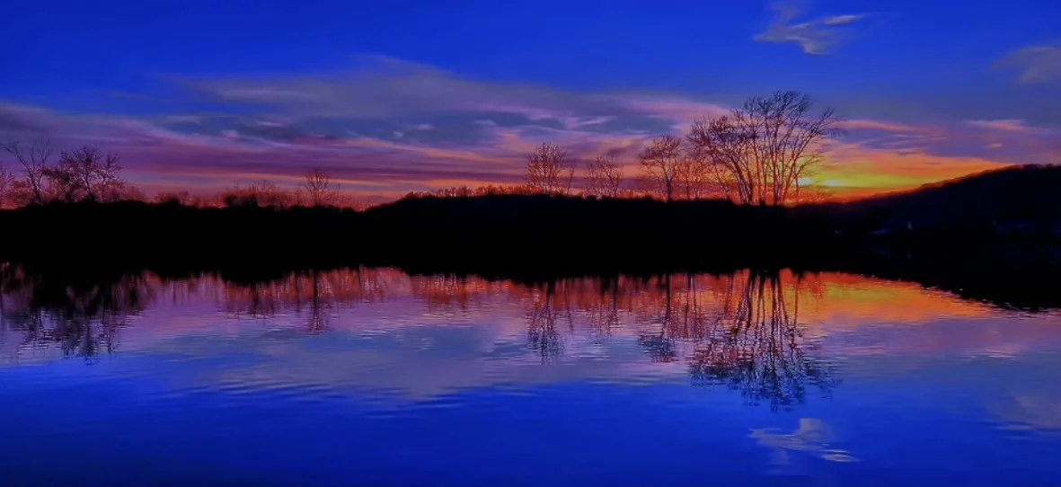 A serene landscape photo of a twilight sky with vibrant orange and purple hues reflected in calm water, silhouetting bare…
