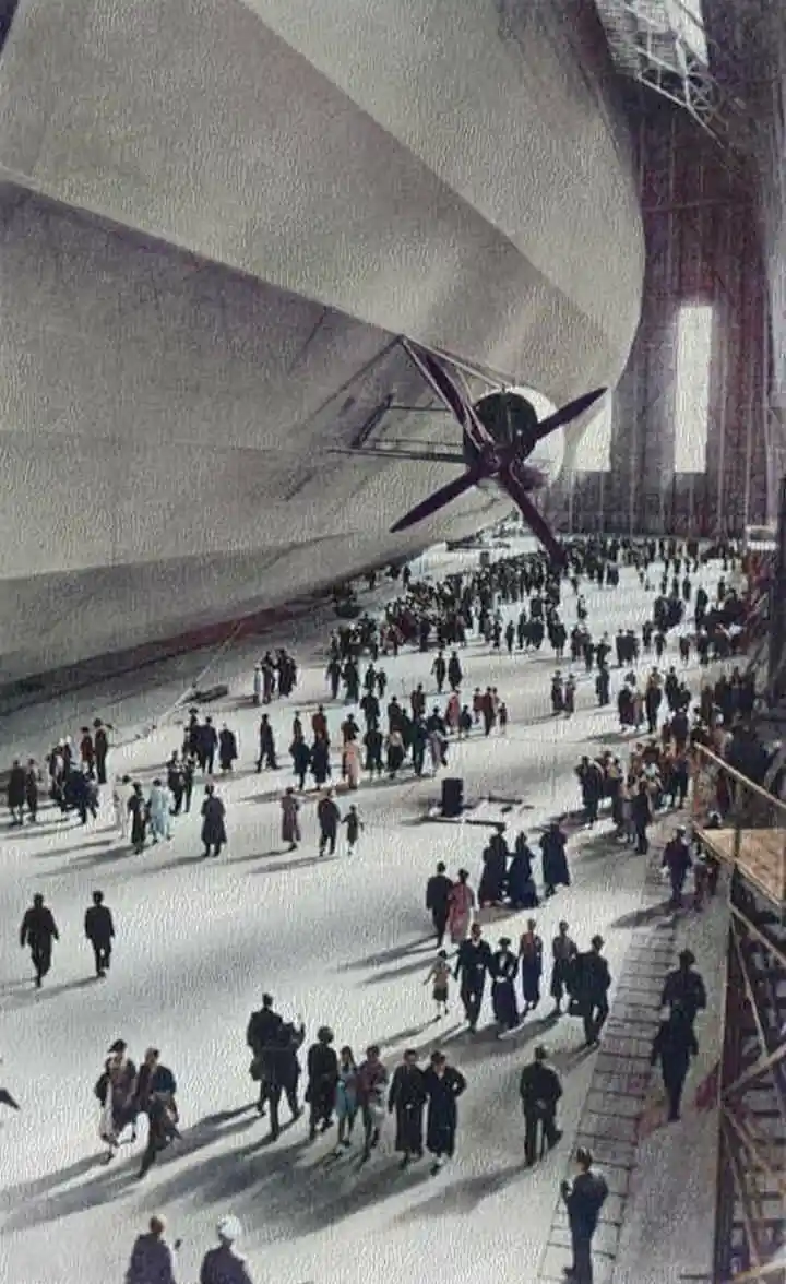 A historical photograph shows a massive zeppelin airship inside a vast hangar, dwarfing the crowds of people walking around…