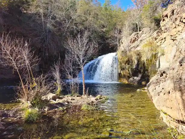A scenic photo of a small waterfall cascading into a clear pool surrounded by rocky cliffs and bare trees under a blue sky…