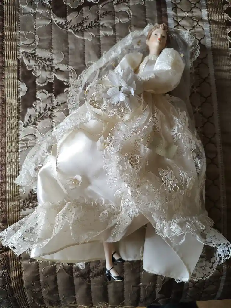A porcelain doll dressed in an elaborate white wedding gown with lace, veil, and floral details lies on a patterned brown…