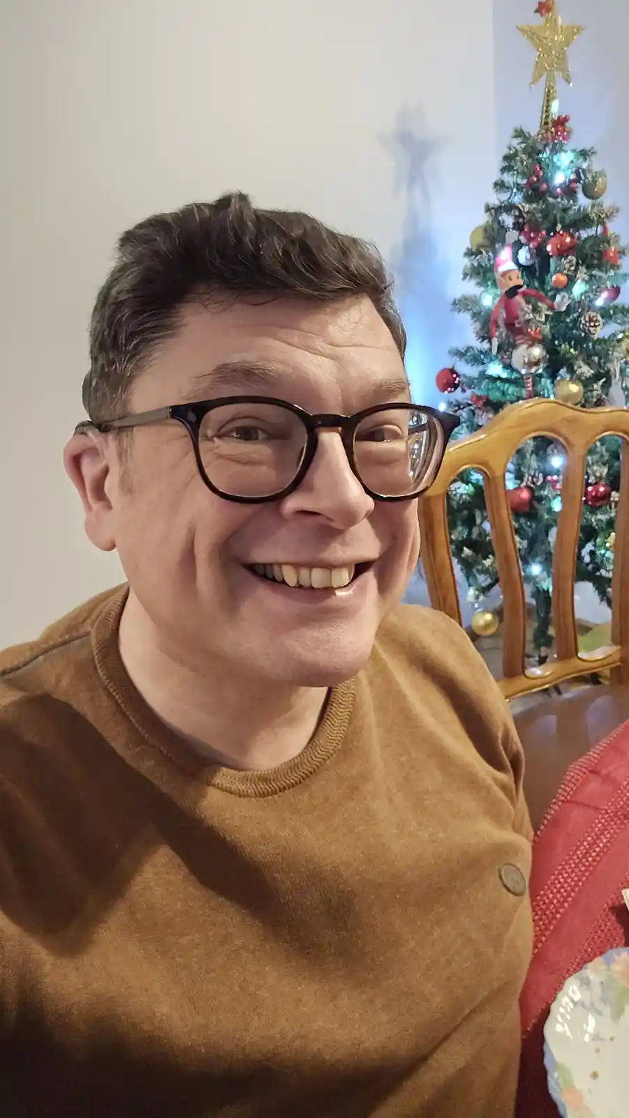 A man with dark hair and black-rimmed glasses smiles broadly at the camera while wearing a brown sweater. Behind him, a…
