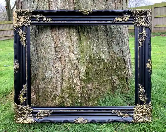A large, ornate black picture frame with gold floral detailing is placed on grass, framing a tree trunk as if it were a…