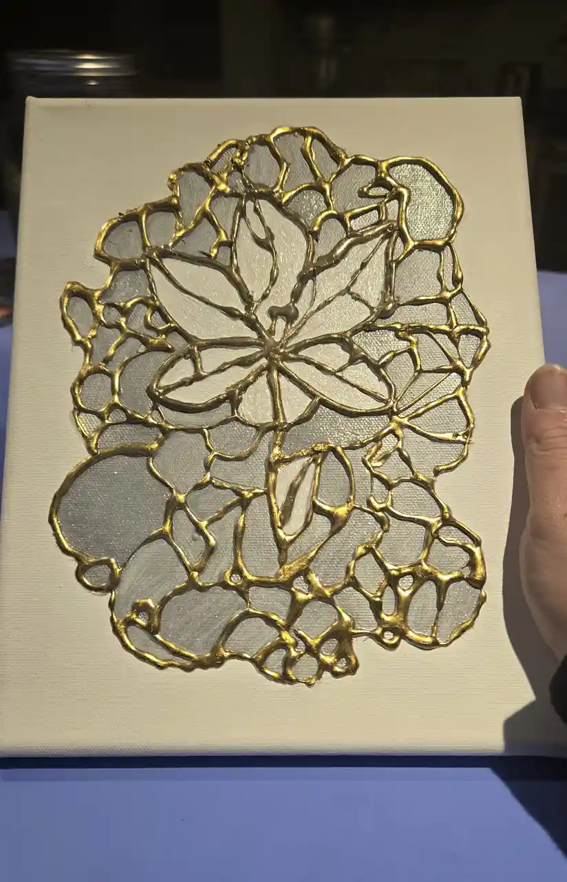 A hand holds up a white canvas displaying a textured, gold-colored abstract artwork resembling a cracked or web-like…