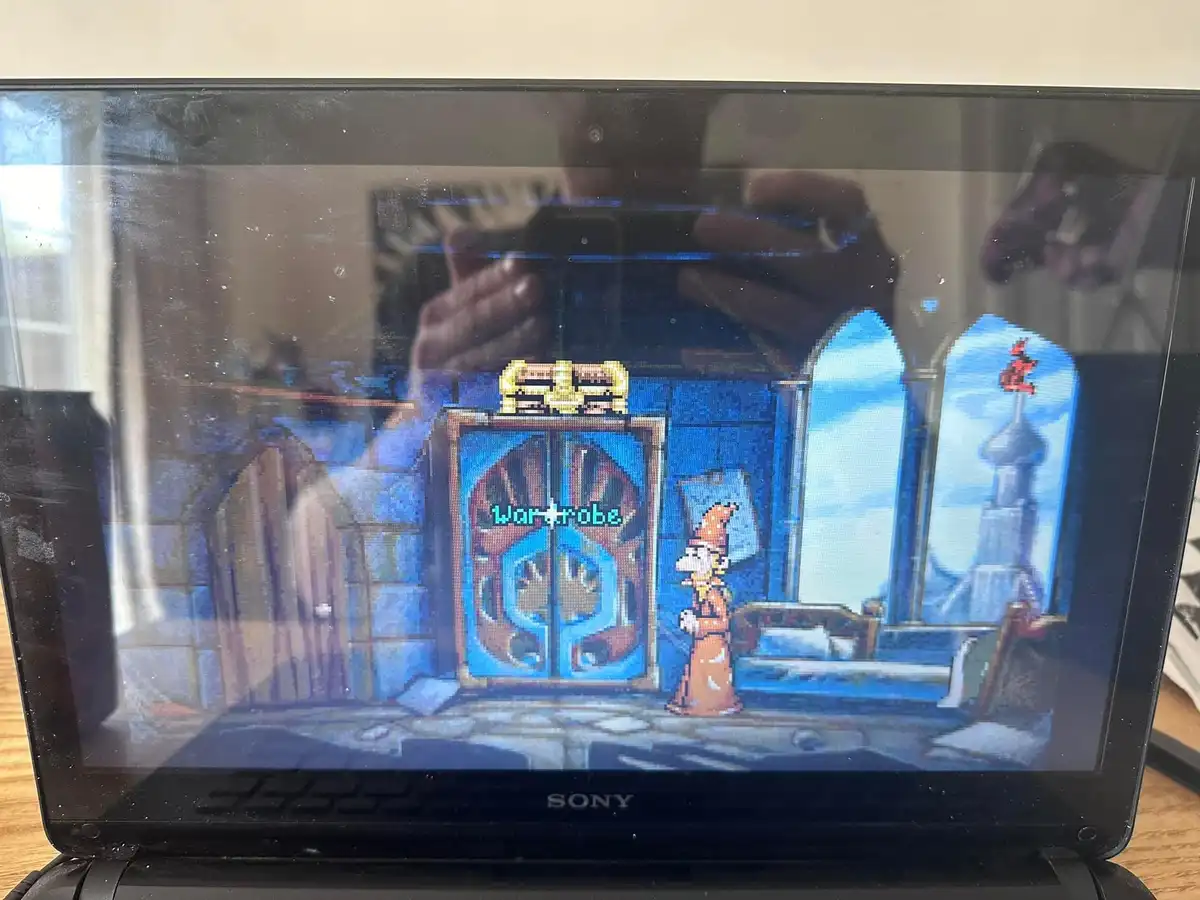 A screenshot of a pixelated video game on a laptop screen shows a character in a brown robe standing before a large blue…