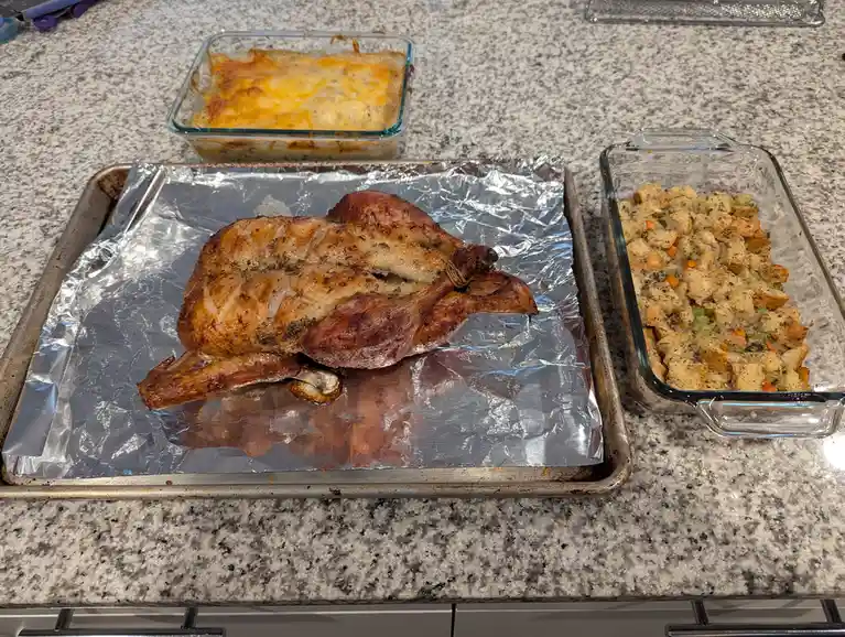 A roasted turkey sits on a foil-lined baking sheet, flanked by glass dishes of stuffing and a cheesy casserole on a granite…