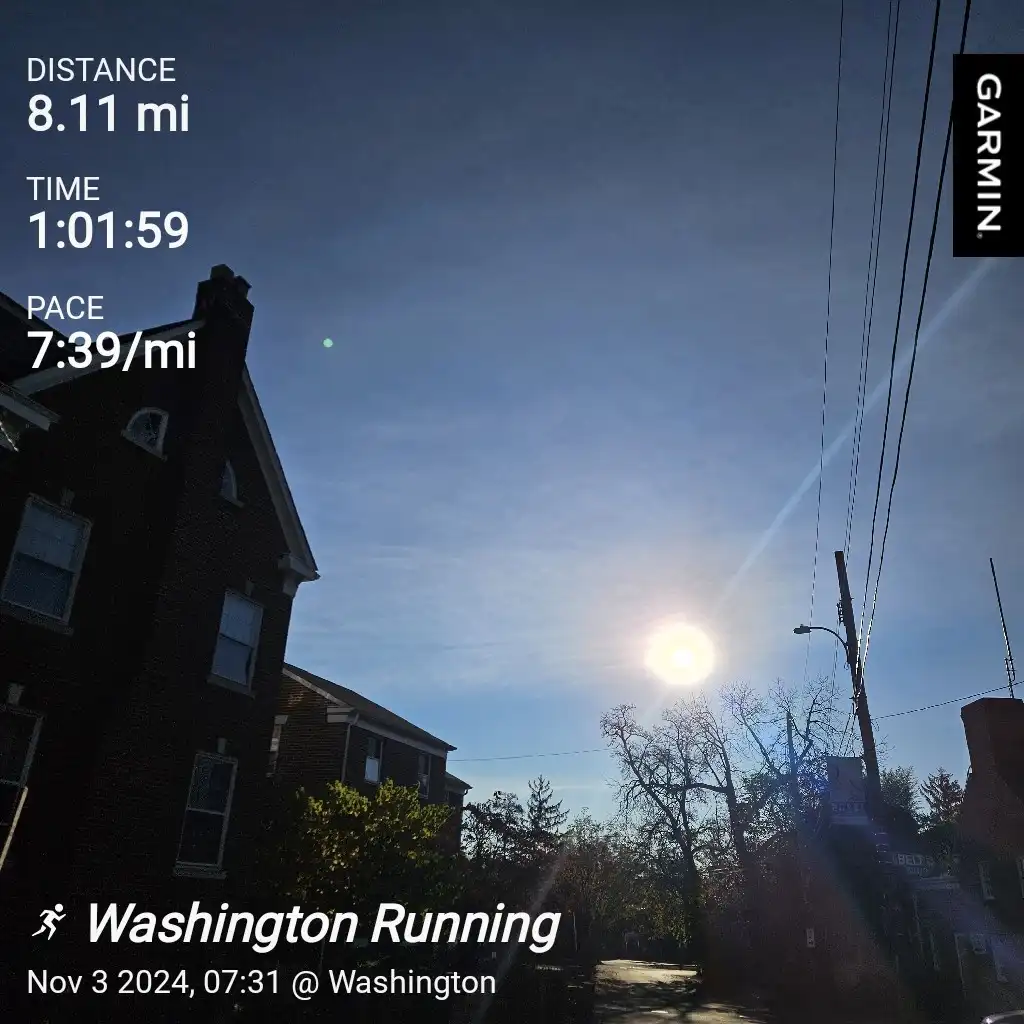 A Garmin running app screenshot shows a bright, sunny morning with a brick house and power lines against a blue sky. The…