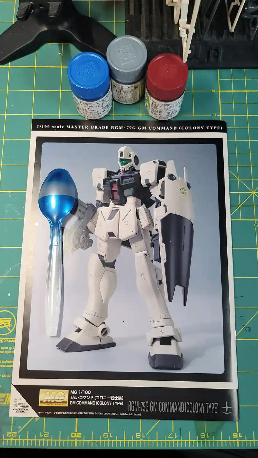 A photo shows a real blue plastic spoon placed next to a printed image of a Gundam model kit box for the RGM-79G GM Command…