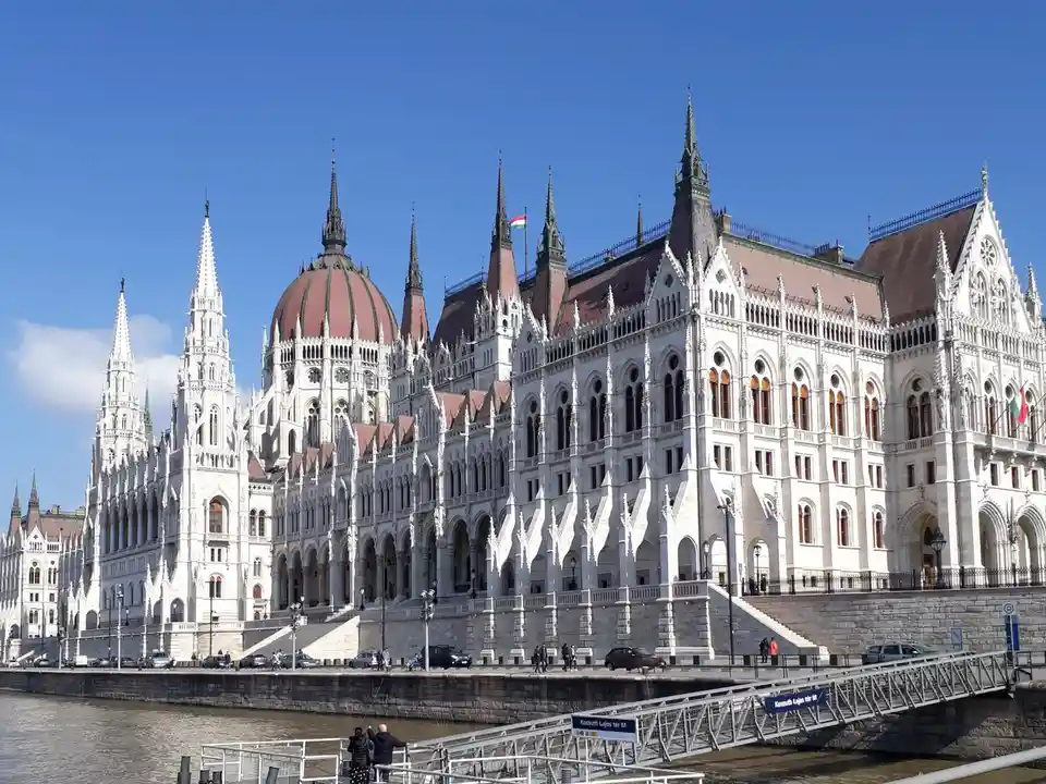 A grand, ornate white Gothic-style building with spires and a red dome stands beside a river under a clear blue sky. A few…
