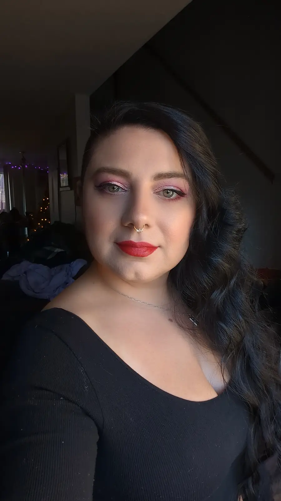 A person with dark wavy hair, wearing a black top, heavy pink eyeshadow, red lipstick, and a nose ring, looks directly at…
