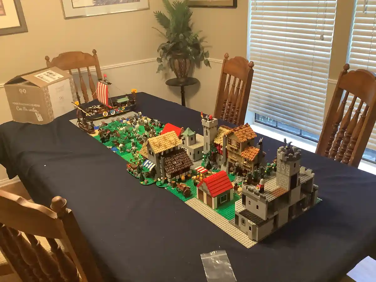 A dining table covered in a dark blue cloth displays an elaborate Lego diorama featuring a medieval village with castles…