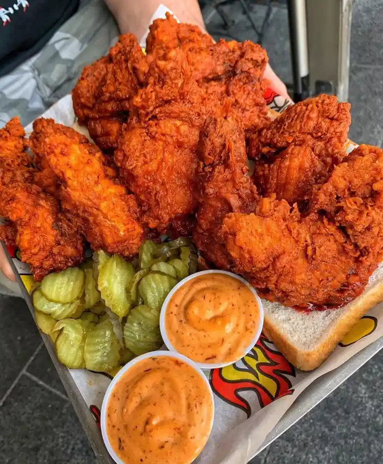 a close-up photo shows a person holding a tray overflowing with crispy, reddish-orange fried chicken pieces, accompanied by…