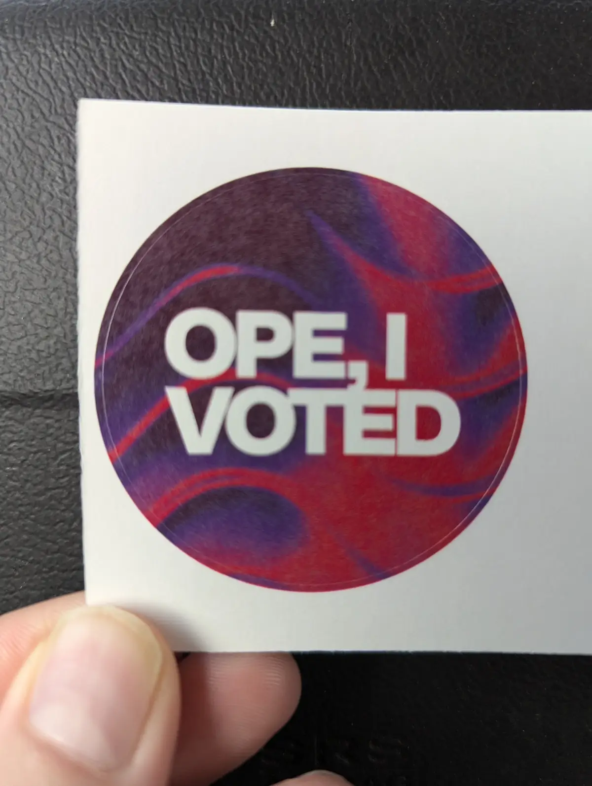 A hand holds a circular sticker with a red and purple marbled background, featuring the white text 'OPE, I VOTED'. The…