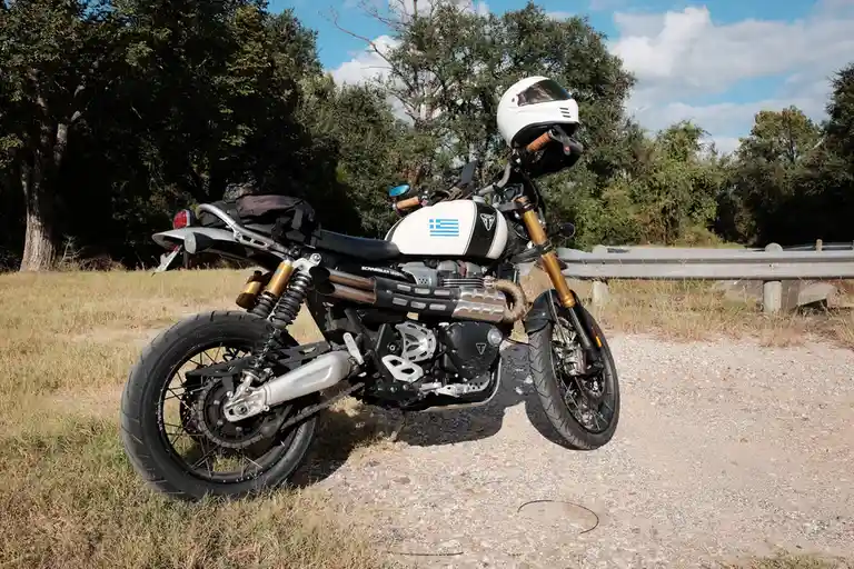 A white and black Triumph Scrambler 1200 motorcycle is parked on a gravel shoulder beside a road with trees and a guardrail…
