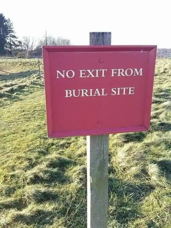 A red rectangular sign with white uppercase text reading 'NO EXIT FROM BURIAL SITE' is mounted on a wooden post in a grassy…