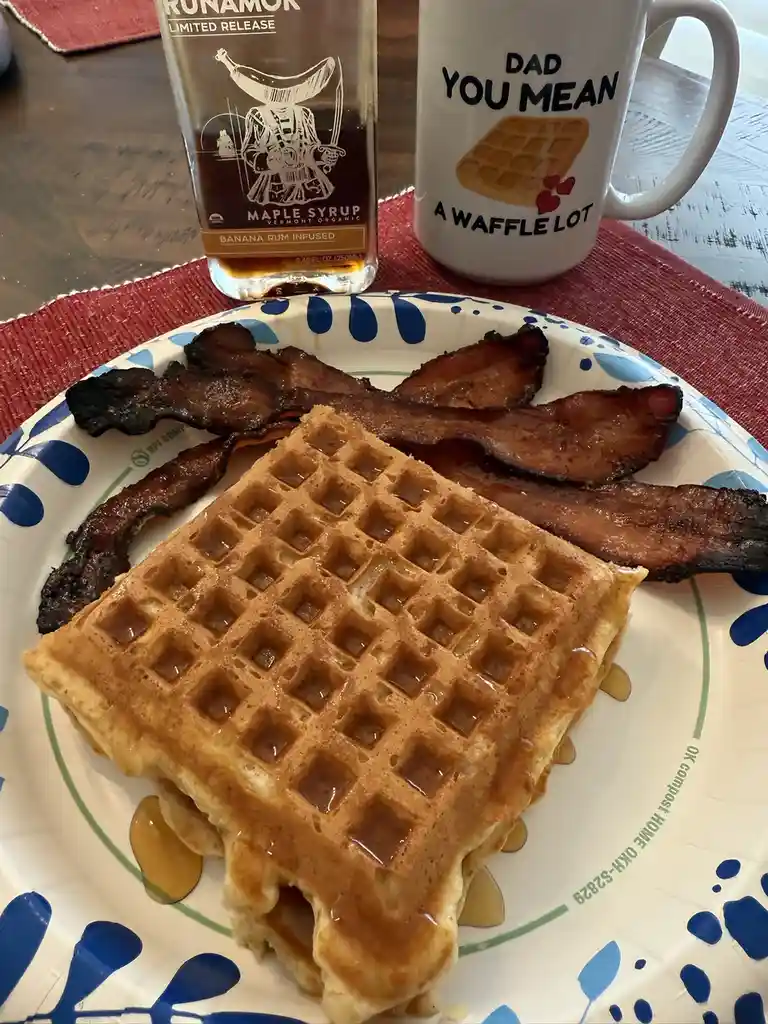 A breakfast scene features a plate of waffles and bacon, with a bottle of banana rum-infused maple syrup and a mug that…