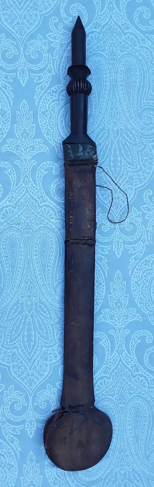 A dark, ornate dagger with a pointed tip and decorative hilt rests vertically against a light blue wallpaper with white…