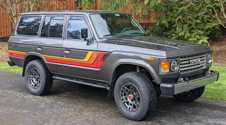 A gray 1980s Toyota Land Cruiser SUV with red and yellow side stripes is parked on a driveway. It has large off-road tires…