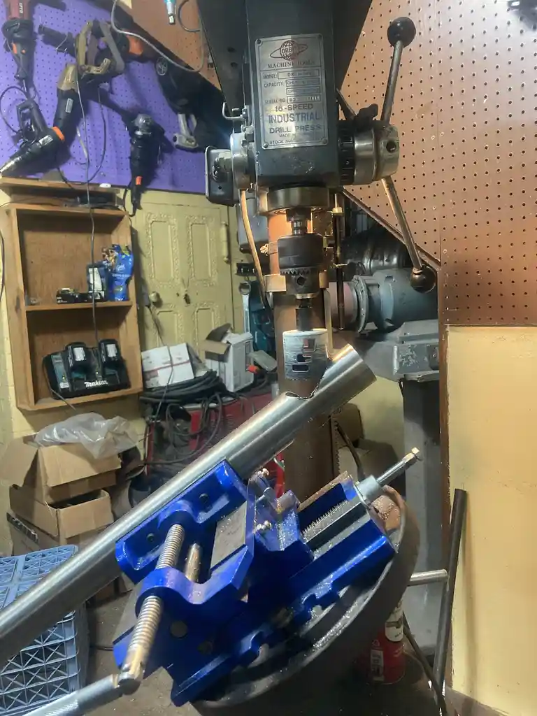 A metal rod is clamped in a blue vise and being drilled by an industrial drill press in a cluttered workshop. The drill bit…