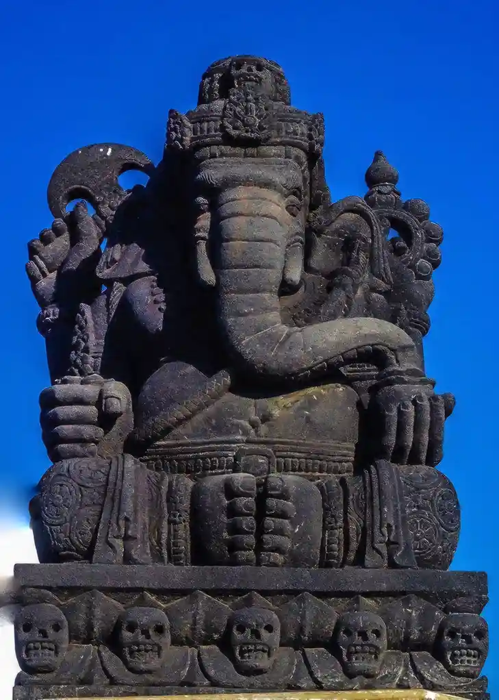 A dark stone statue of the Hindu deity Ganesha, depicted with an elephant head and multiple arms, sits against a bright blue…