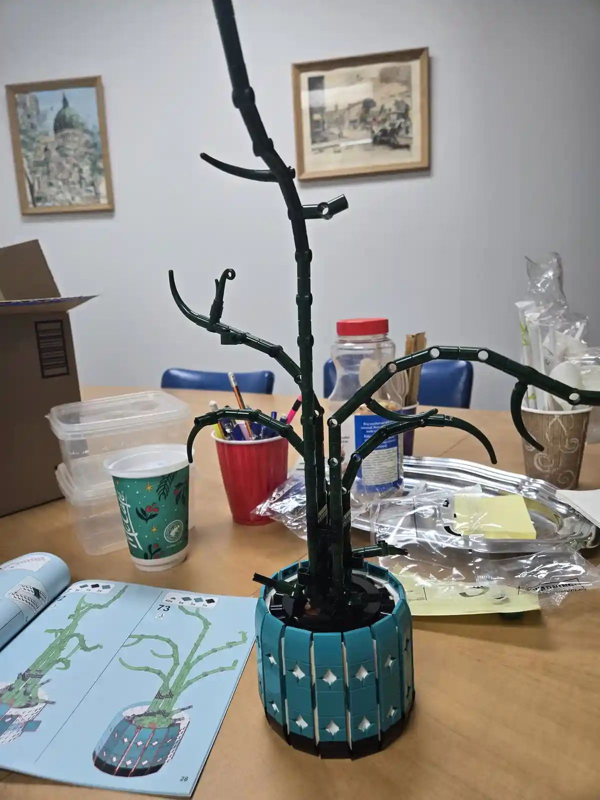 A partially assembled green LEGO tree model sits on a wooden table next to its instruction manual, which shows a completed…