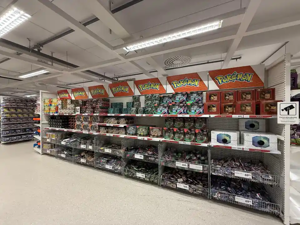 A wide shot of a well-lit toy store aisle filled with Pokémon merchandise, including action figures, cards, and collectibles…