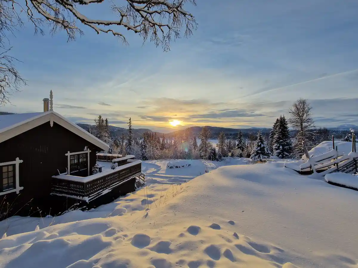 A serene winter scene shows a dark wooden cabin with snow-covered roof and balcony, set against a snowy landscape with…
