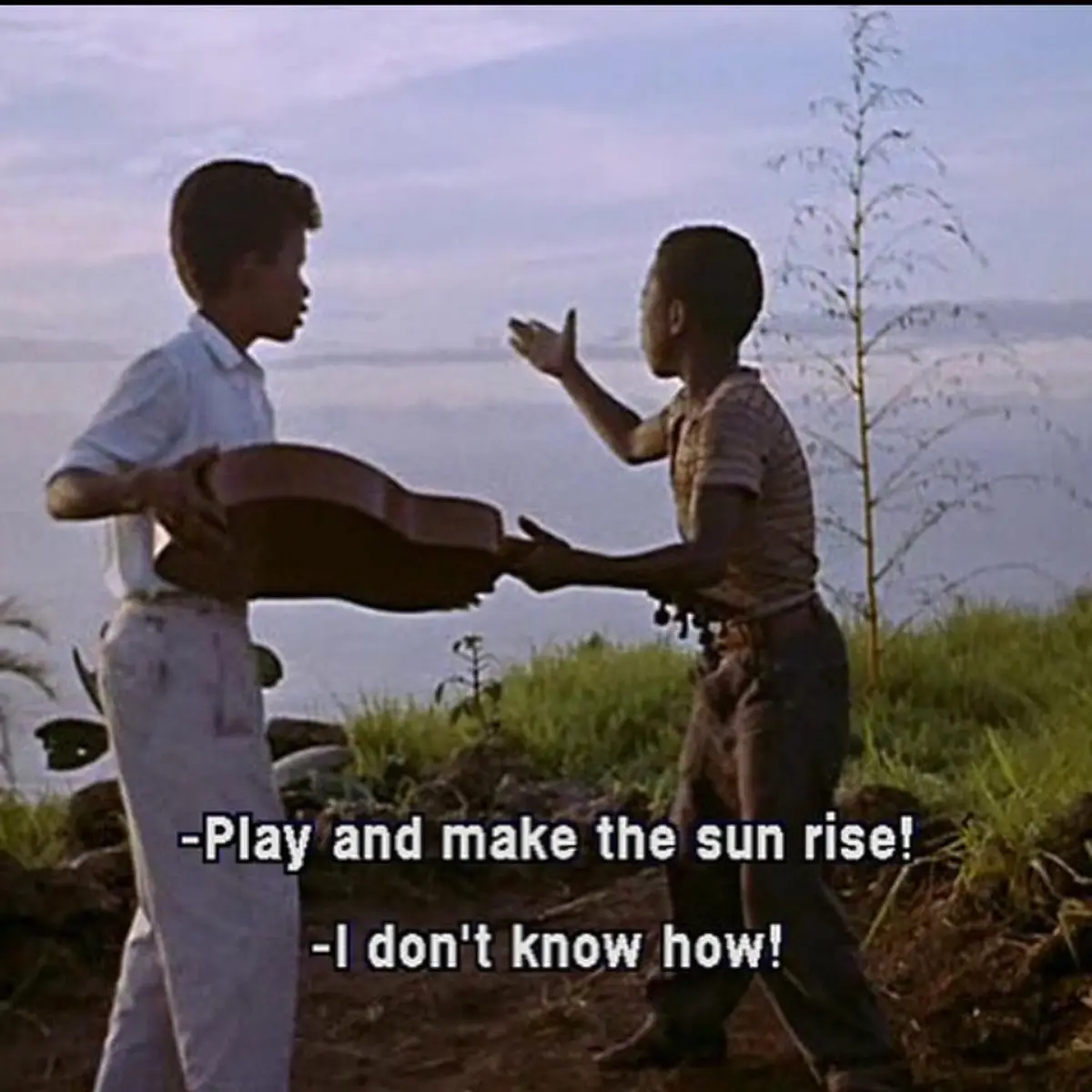 Two young boys stand outdoors on a grassy hillside, one holding a guitar while the other gestures toward him. The overlaid…