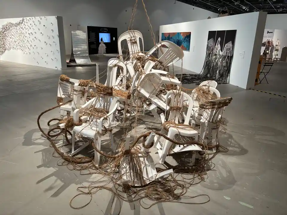 A large art installation in a gallery features numerous white plastic chairs bound together with thick brown rope, suspended…