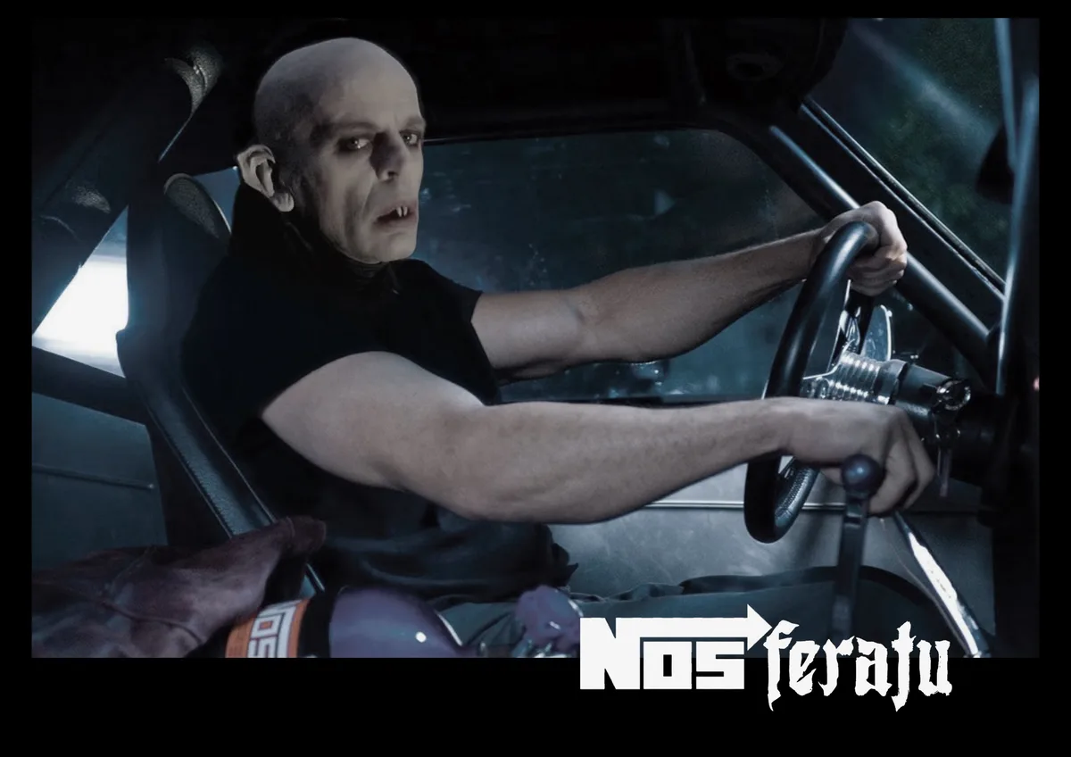 A bald, pale vampire with fangs grips a car's steering wheel and gear shift, looking intensely forward. The image has a…