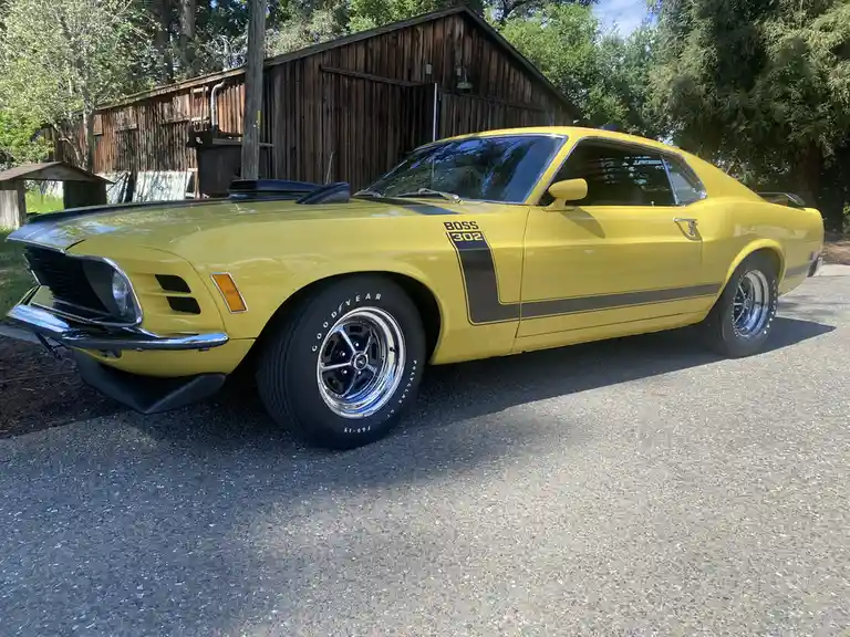 A bright yellow 1970 Ford Mustang Boss 302 is parked on asphalt in front of a wooden barn, featuring black racing stripes…