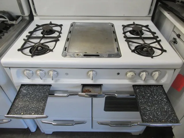 A white vintage-style gas stove with four burners and two oven doors, both of which are open. The oven doors have speckled…