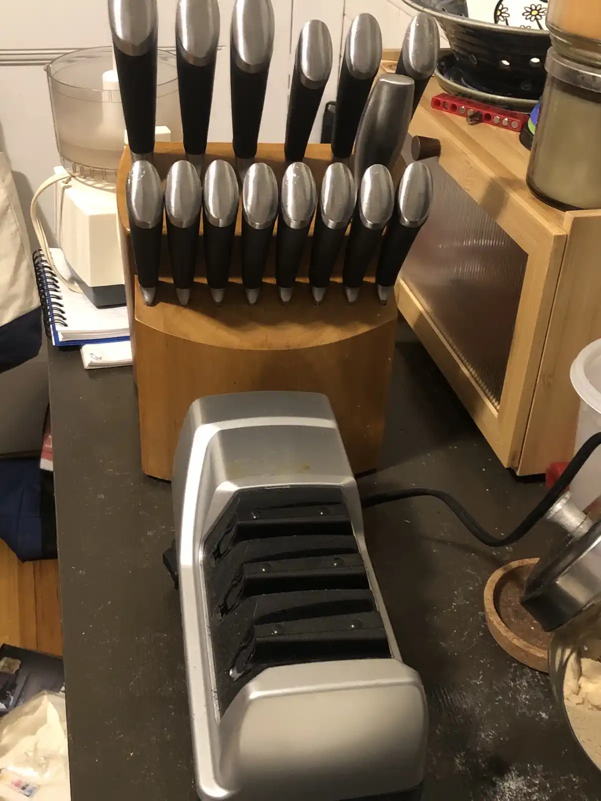 A wooden knife block filled with numerous knives stands behind a silver electric knife sharpener on a dark countertop. The…