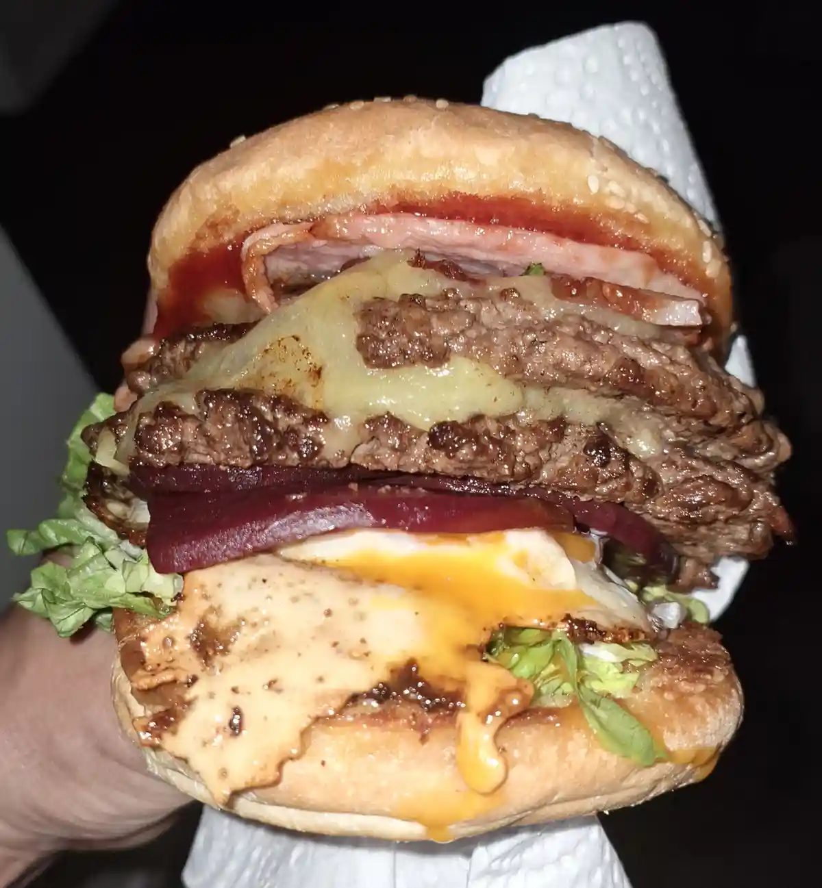 A hand holds a massive, towering burger with multiple beef patties, melted cheese, bacon, onions, lettuce, and sauce, all…