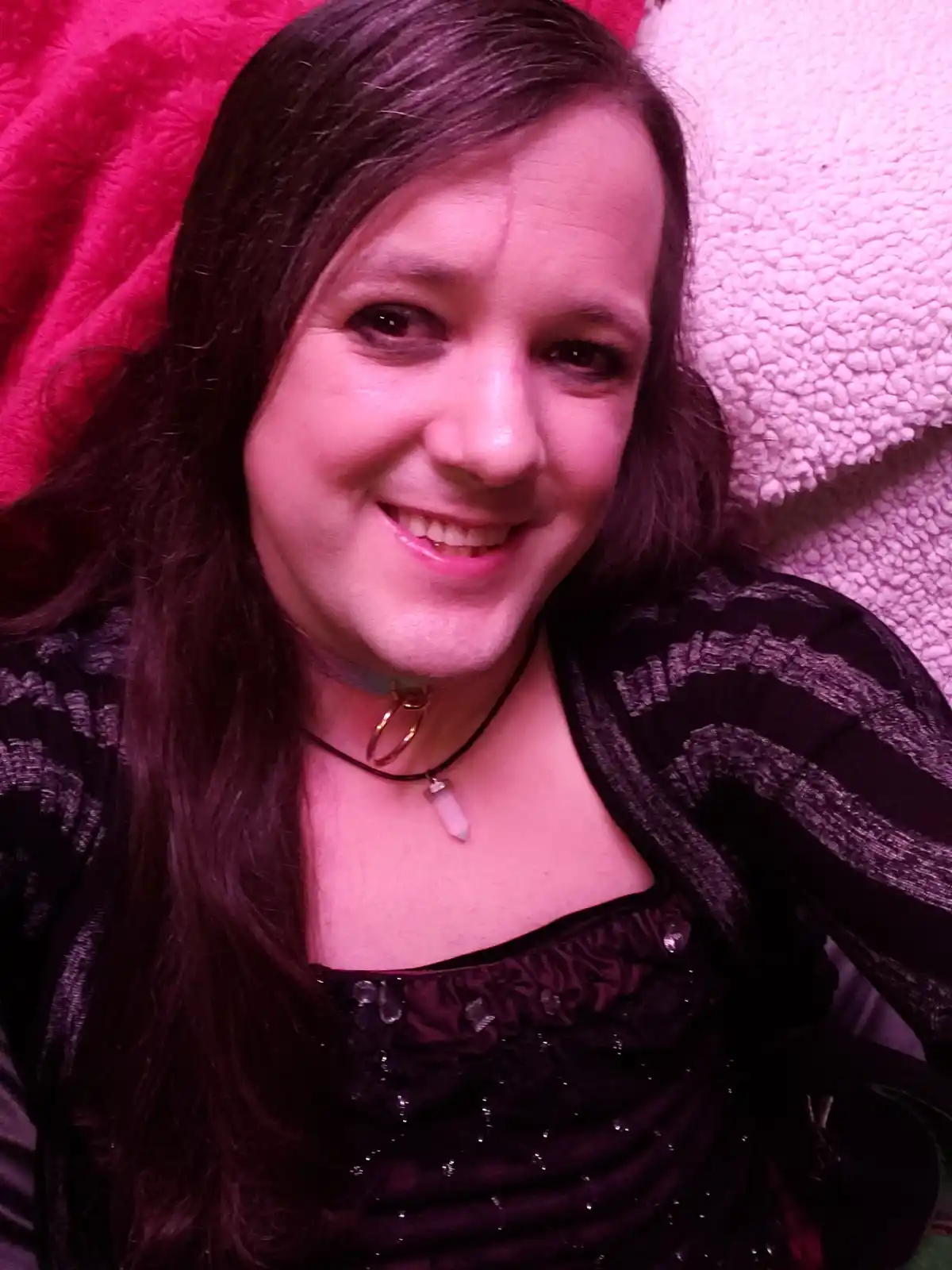 A person with long dark hair smiles at the camera while lying on a pink and white textured blanket, wearing a black top with…