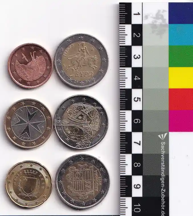 Six euro coins from different countries are arranged on a white surface next to a ruler and color calibration chart. The…