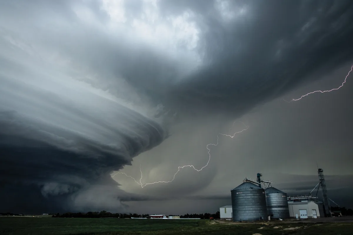 A dramatic photograph shows a massive, dark storm cloud looming over a rural landscape with two large metal grain silos and…