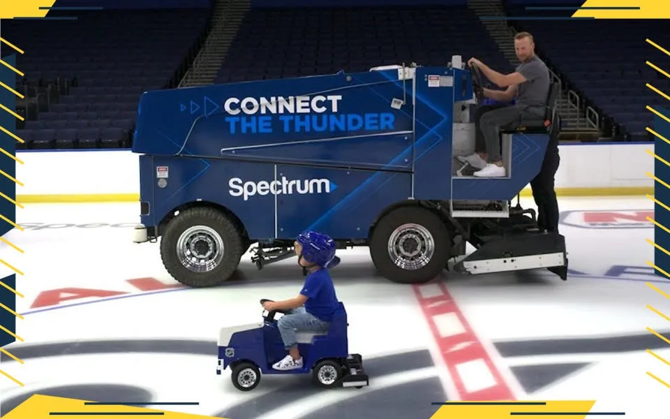 A man drives a large blue ice resurfacer on an ice rink, while a small child in a blue helmet drives a miniature replica of…