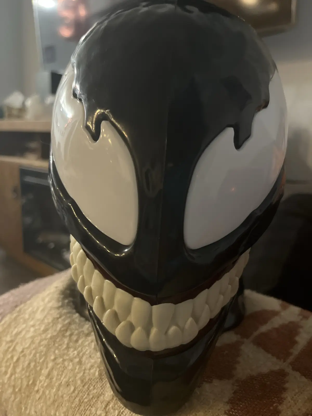 A glossy black and white Venom mask with large white eyes and sharp teeth is shown in a close-up, resting on a textured…