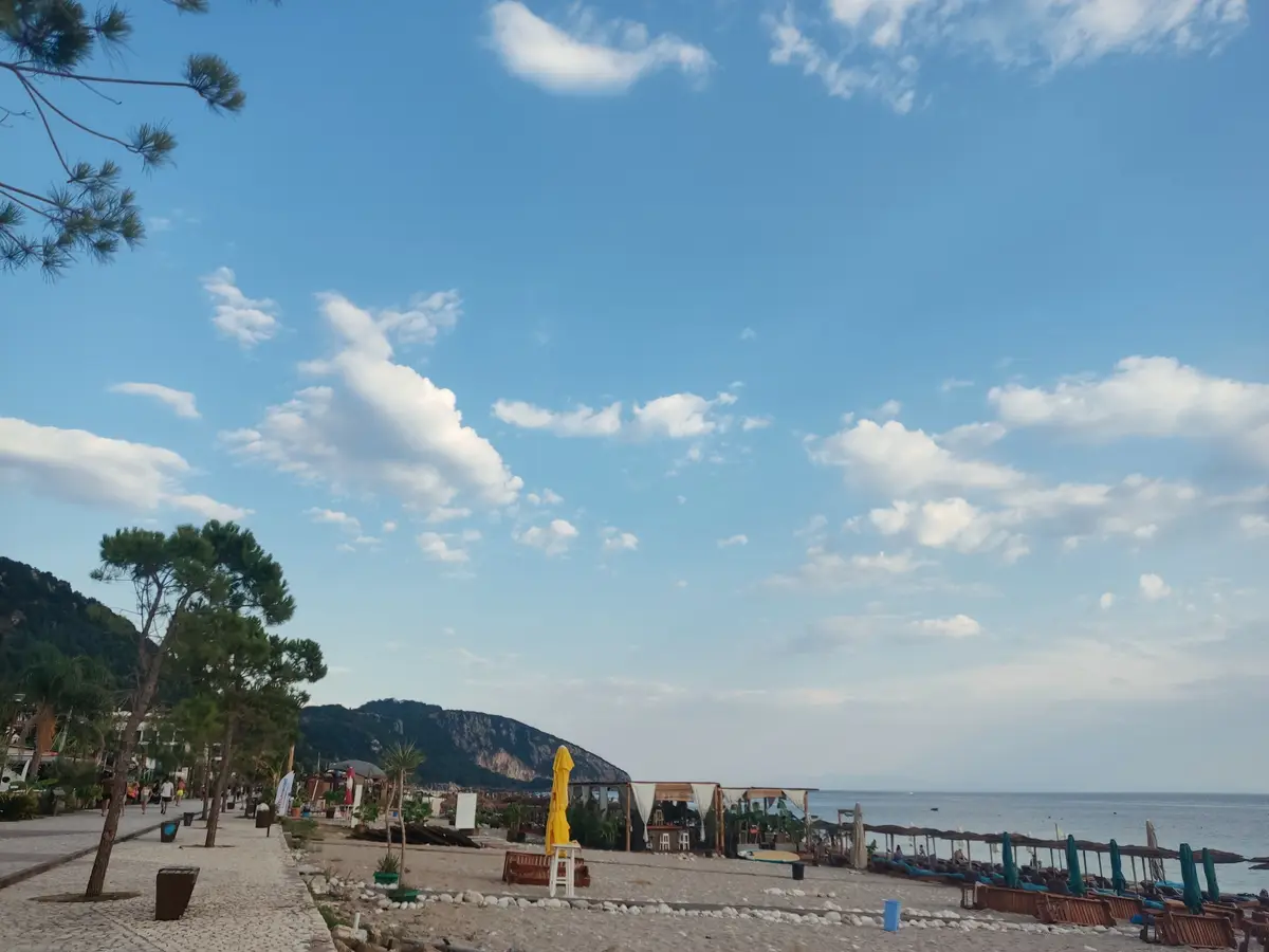 A scenic beach photo with a clear blue sky, scattered clouds, pine trees, and a coastal path leading to a beach with lounge…