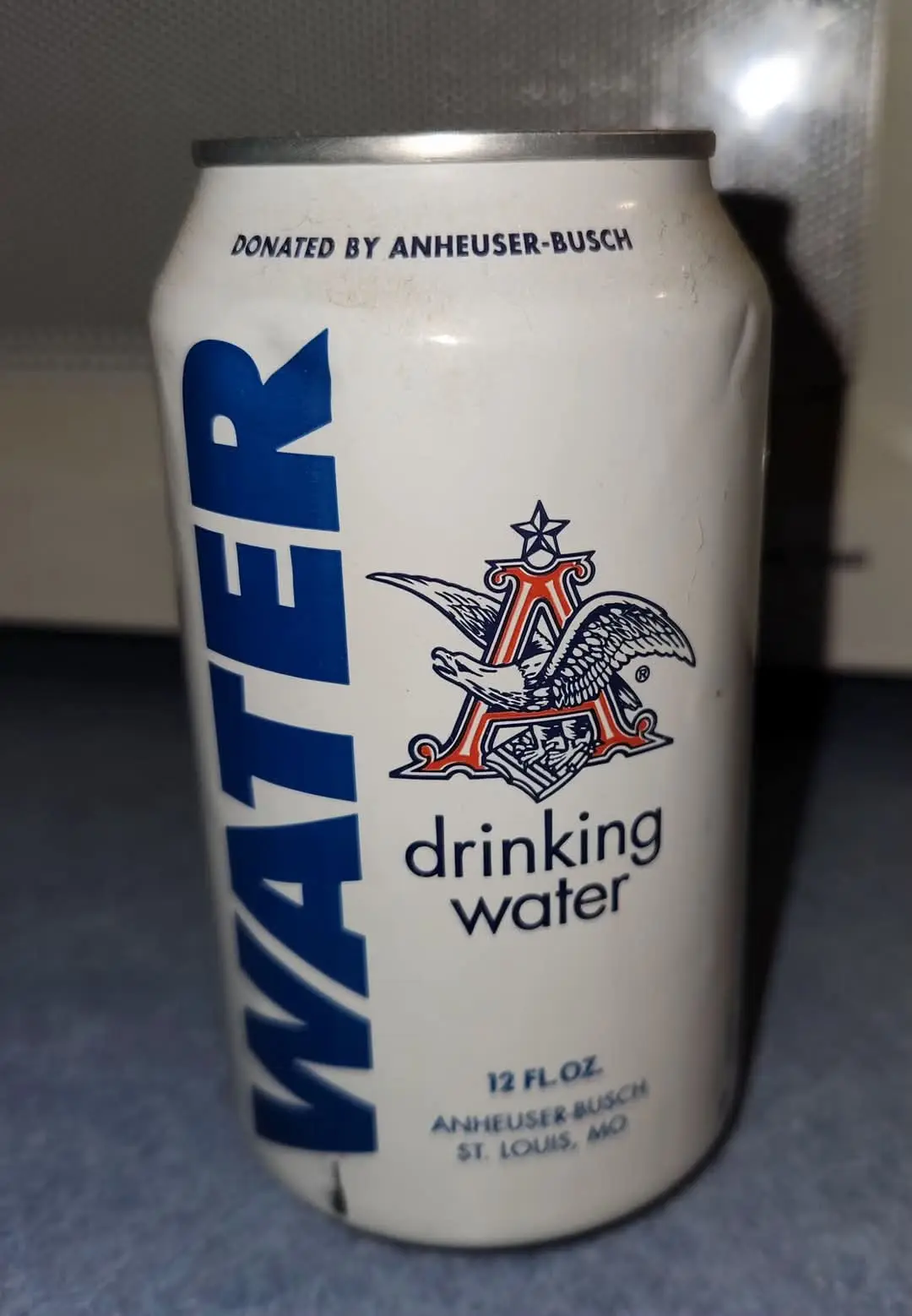 A white aluminum can labeled 'WATER' vertically in blue, featuring the Anheuser-Busch eagle logo and text 'drinking water'…