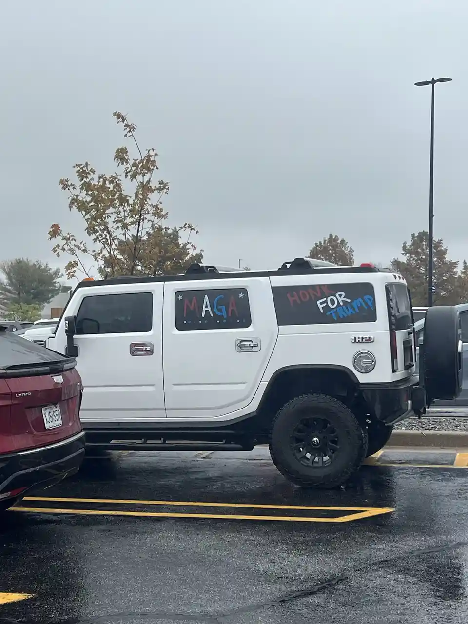 A white Hummer H2 parked in a wet parking lot has 'MAGA' written in colorful letters on its rear passenger window and 'HONK…