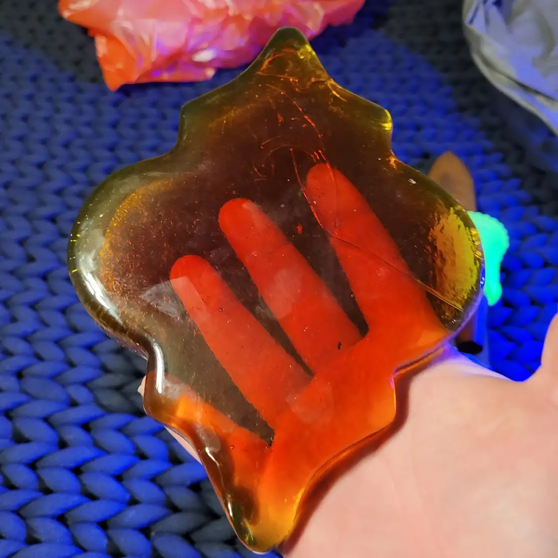A hand is holding a translucent, amber-colored resin object shaped like a leaf, with a clear handprint impression inside…