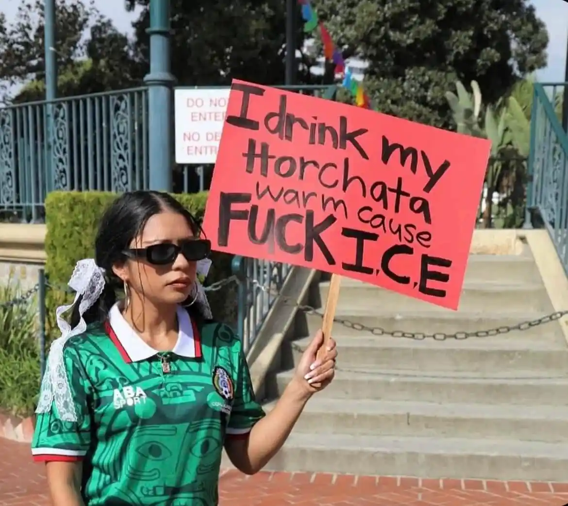 A woman wearing a green Mexican-themed jersey and sunglasses holds a red sign with black handwritten text that reads 'I…