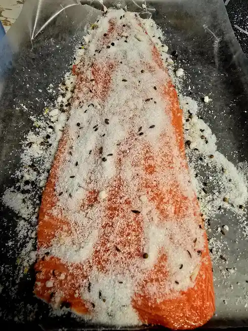 A raw salmon fillet lies on aluminum foil, heavily coated with a thick layer of white granular salt and scattered dark…