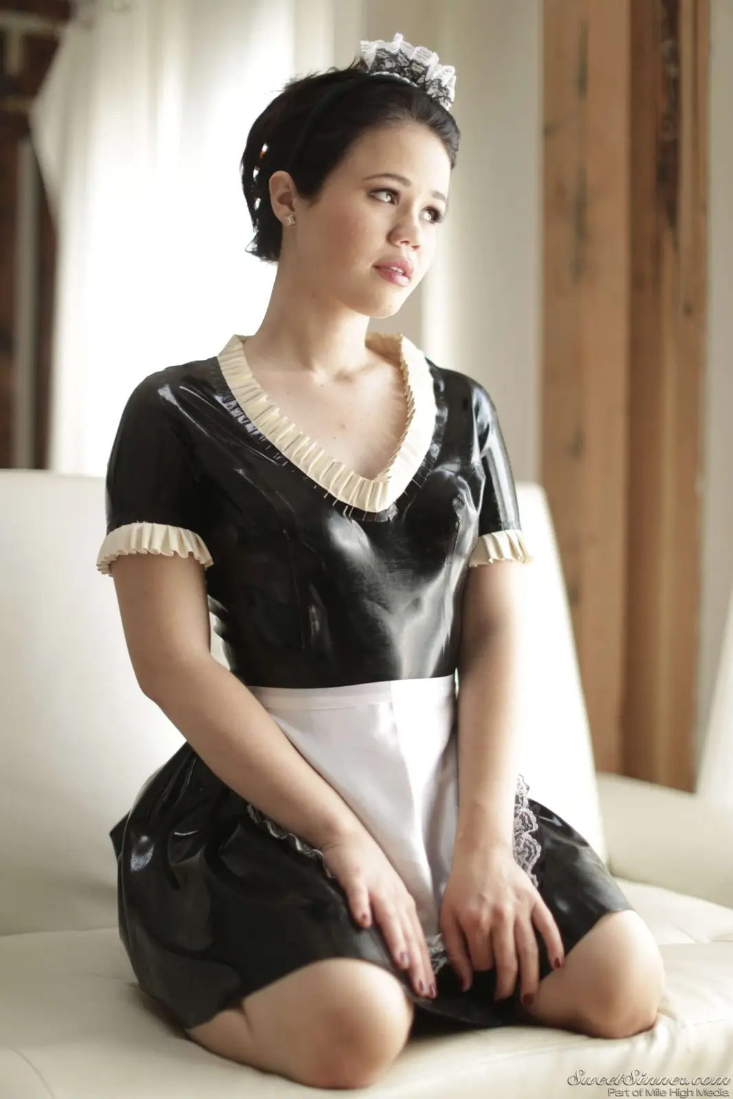 A woman in a black latex maid outfit with white lace trim and a white apron sits on a white couch, looking off to the side…
