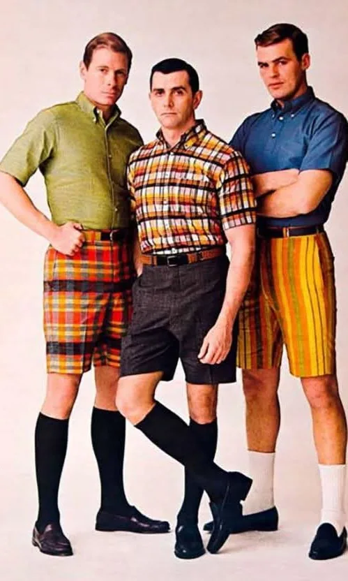 Three men in vintage 1960s-style outfits pose seriously; two wear plaid shorts with knee-high socks and loafers, while the…