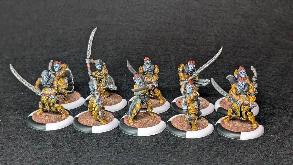 Six painted miniature fantasy warriors, each with blue skin, red hair, and gold armor, stand on circular bases with…